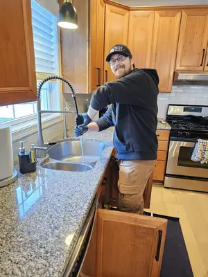 A man wearing a cap and gloves washes dishes at a kitchen sink with wooden cabinets and stainless steel appliances.