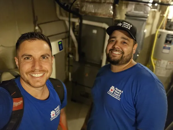 Two men in blue shirts with "Andy's Statewide Heating & Cooling" logos stand in a basement near HVAC equipment, smiling at the camera.