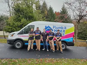 Four workers in matching uniforms stand in front of a company van labeled "Plumb it Heat it" parked on a driveway with trees in the background.