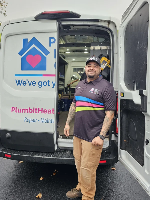A repair technician stands smiling by an open service van with the company logo, wearing a branded shirt and cap, on a wet asphalt surface.