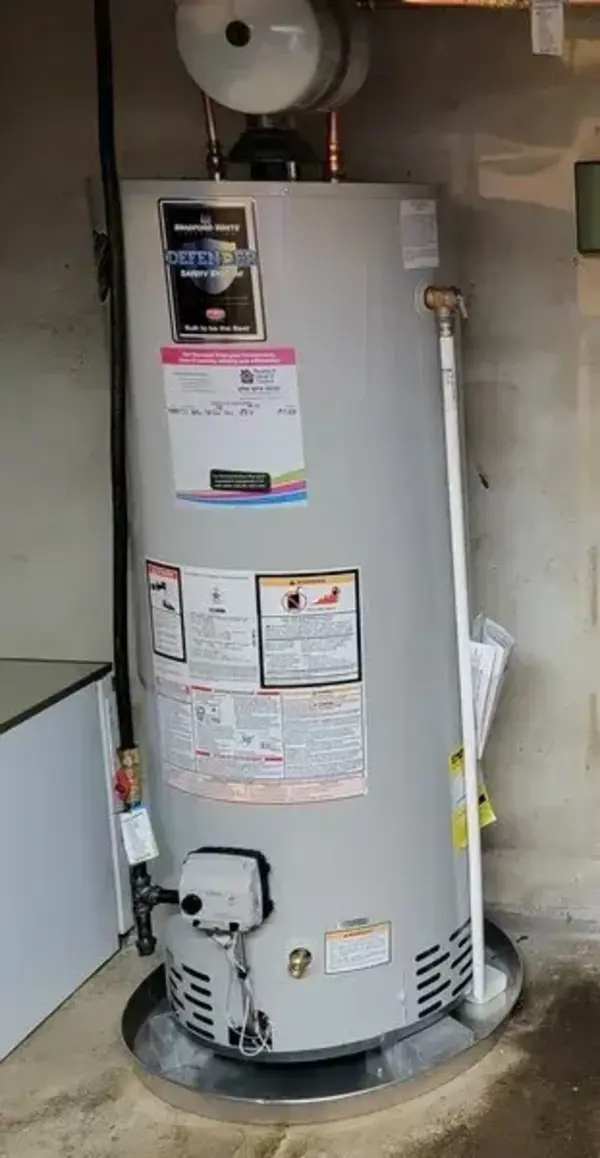 Tall cylindrical water heater installed in a basement, with labels and instructions on the front, and pipes connected at the top and side.