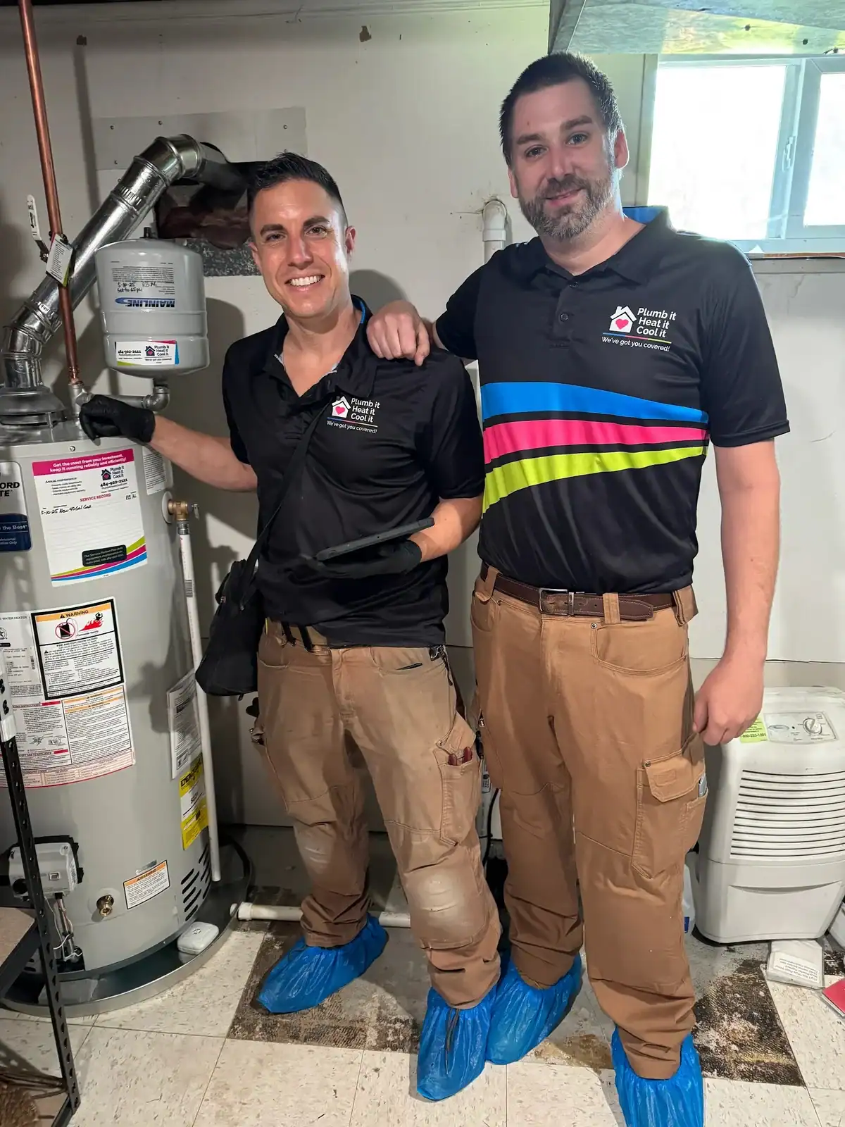 Two plumbers in black shirts, brown pants, and blue shoe covers stand next to a water heater in a basement, ready to handle any Water Heater Repair needs.