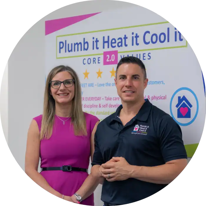 Two people stand before a sign that showcases the "Plumb it Heat it Cool it CORE 2.0 VALUES." The woman is in a pink dress with glasses, while the man sports a black Plumb it Heat it Cool it company polo shirt.