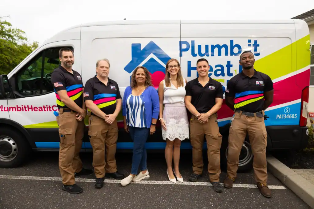 Five people stand in front of a Plumb-It Heat It Cool It Health Care van, with two women in the center and three men in work uniforms on either side.