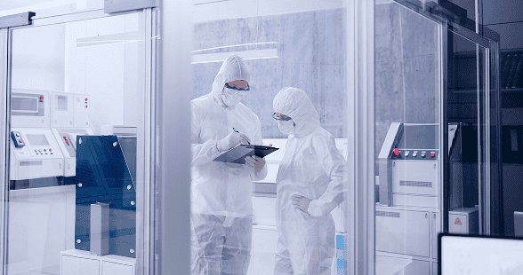 Two technicians in full cleanroom protective suits and masks standing inside a sterile lab environment reviewing information on a tablet.