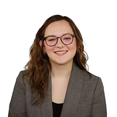 Headshot of a woman with curly hair wearing glasses and a gray blazer against a white background.