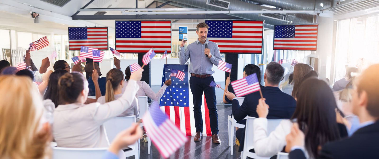 A man speaking at a political event with attendees waving American flags in a conference room.