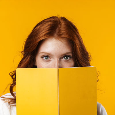 A woman with red hair holding a yellow book in front of her face against a yellow background.