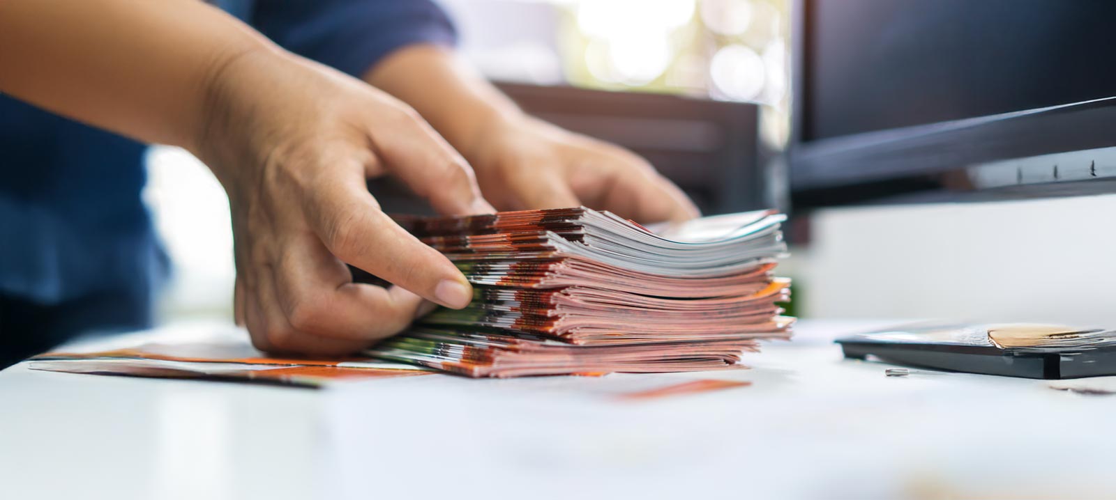 A person organizing a stack of printed leaflets on a desk next to a computer.