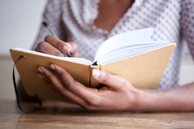 A person holding a hard cover journal with blank pages, writing with a pen.
