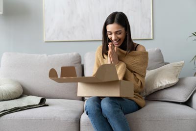 A woman sitting on a couch, looking excited while opening a brown cardboard box.