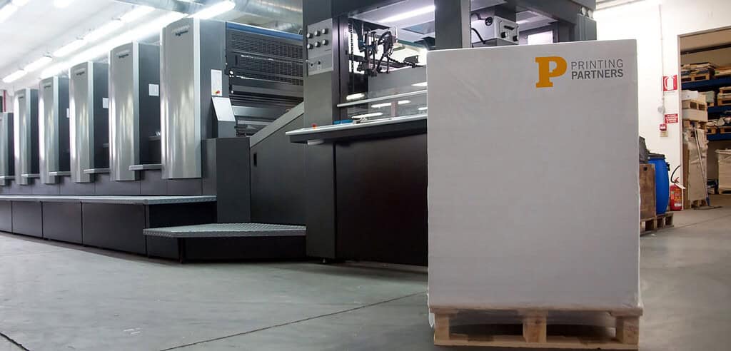 A stack of printed materials on a pallet in front of printing machinery at Printing Partners.