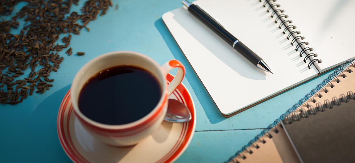 A spiral bound journal next to a cup of coffee on a blue surface.