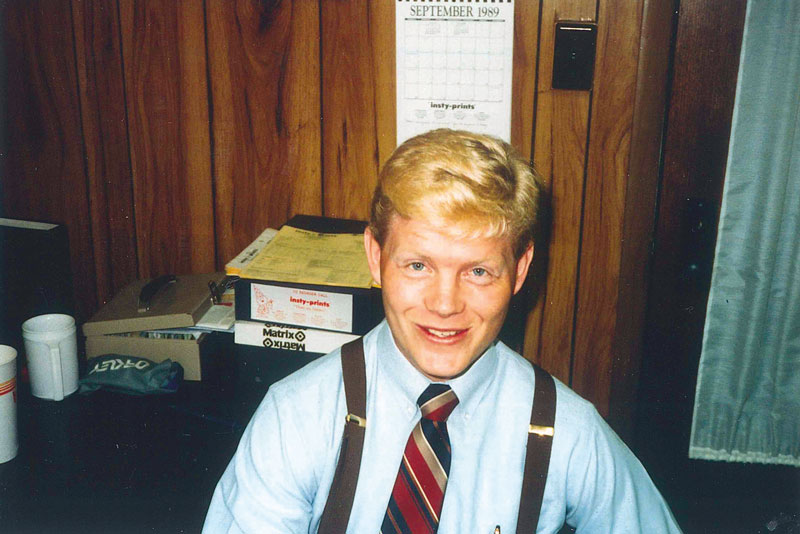 A smiling man with blonde hair wearing a blue shirt and a striped tie, seated at a desk.