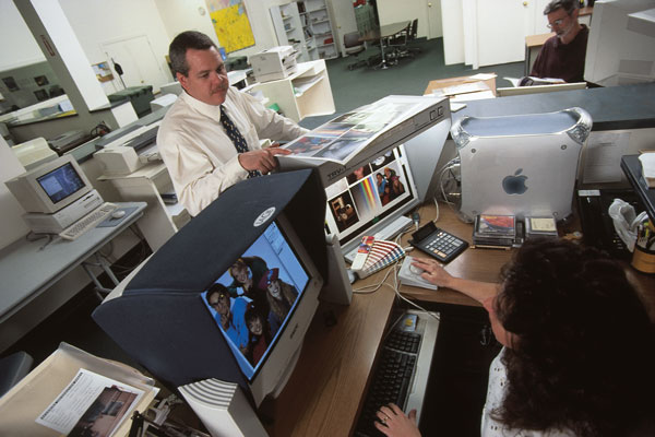 An office scene featuring two individuals working at computers and a third person holding printed materials.