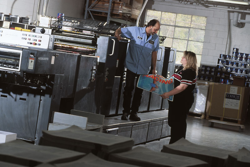 Two individuals interacting near a printing press in a production facility.