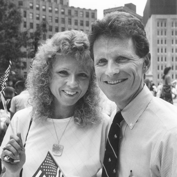 Black and white image of two smiling individuals holding small American flags.