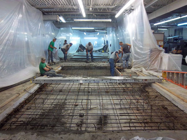 Workers installing a cement pad at a construction site with exposed rebar and plastic sheeting.