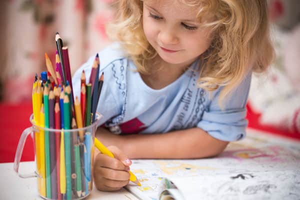 A young child coloring with a yellow pencil, surrounded by a jar of colored pencils.