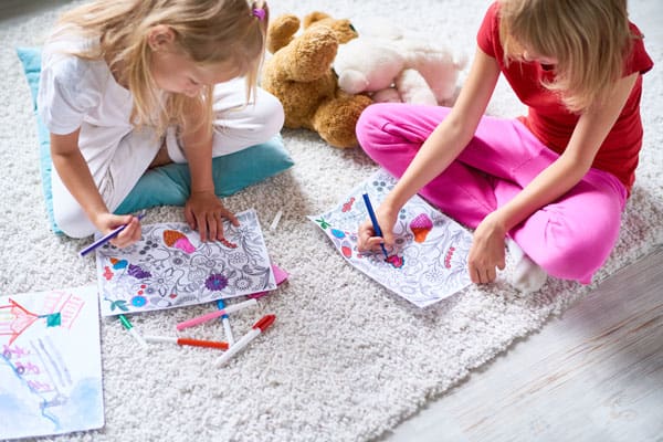 Two girls sitting on a carpet, coloring on sheets of paper with markers.