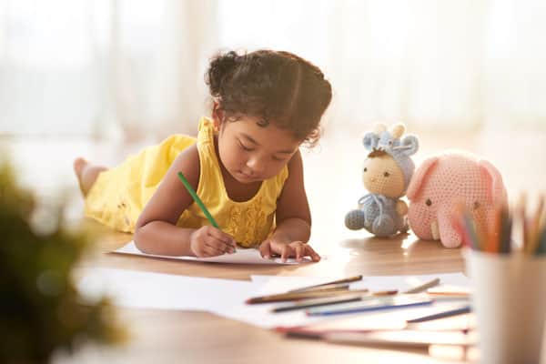 A young child in a yellow dress coloring on a sheet of paper with crayons and stuffed toys nearby.