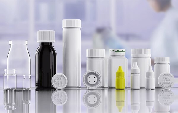 A variety of pharmaceutical bottles and containers, including pill bottles, dropper bottles, and pill organizers, arranged on a reflective surface in a laboratory setting.