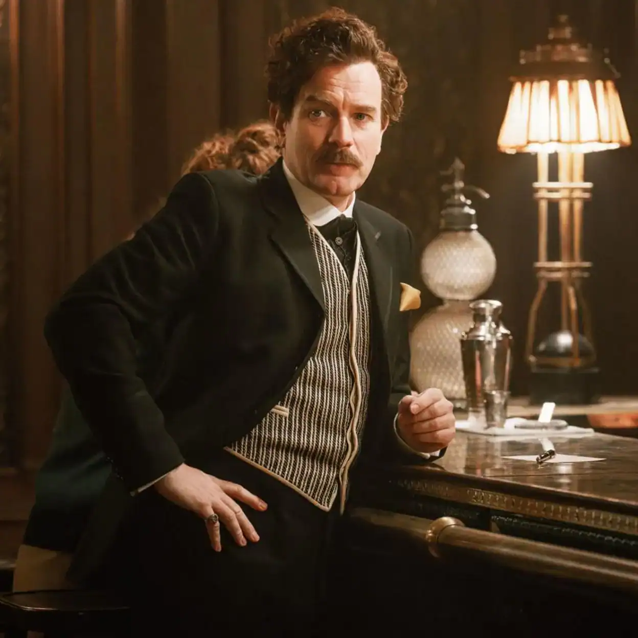 Un hombre con bigote y estilo clásico, vestido con traje elegante, en un entorno vintage con lámparas de luz cálida, en una escena que evoca películas de época en un bar sofisticado.