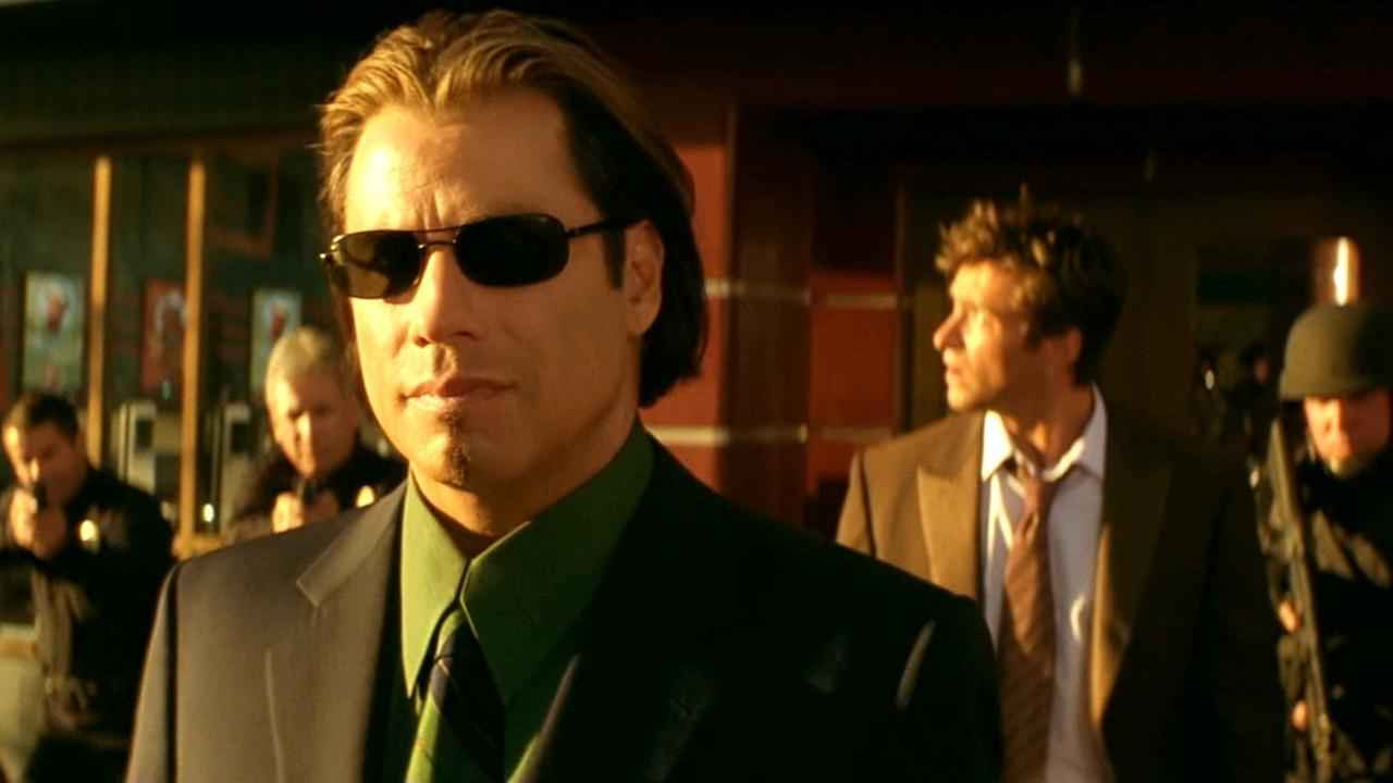 Un hombre con gafas de sol calvo y traje negro en una escena de película, en un ambiente policial o de investigación en una ciudad moderna.