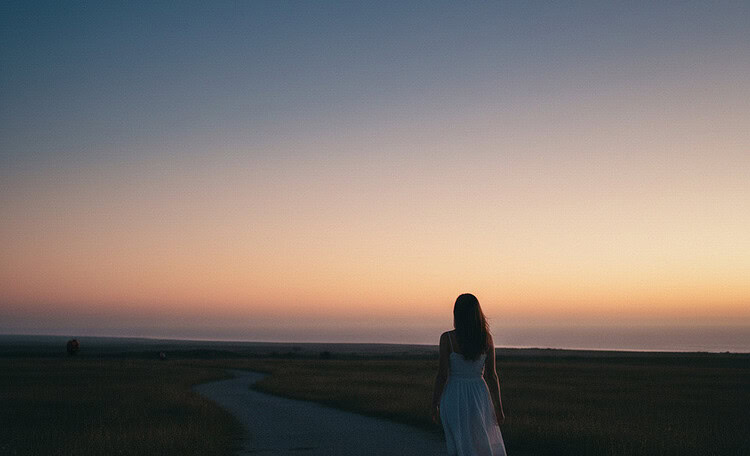Woman in a white dress walks along a winding path at dusk toward a colorful sunset over open fields.