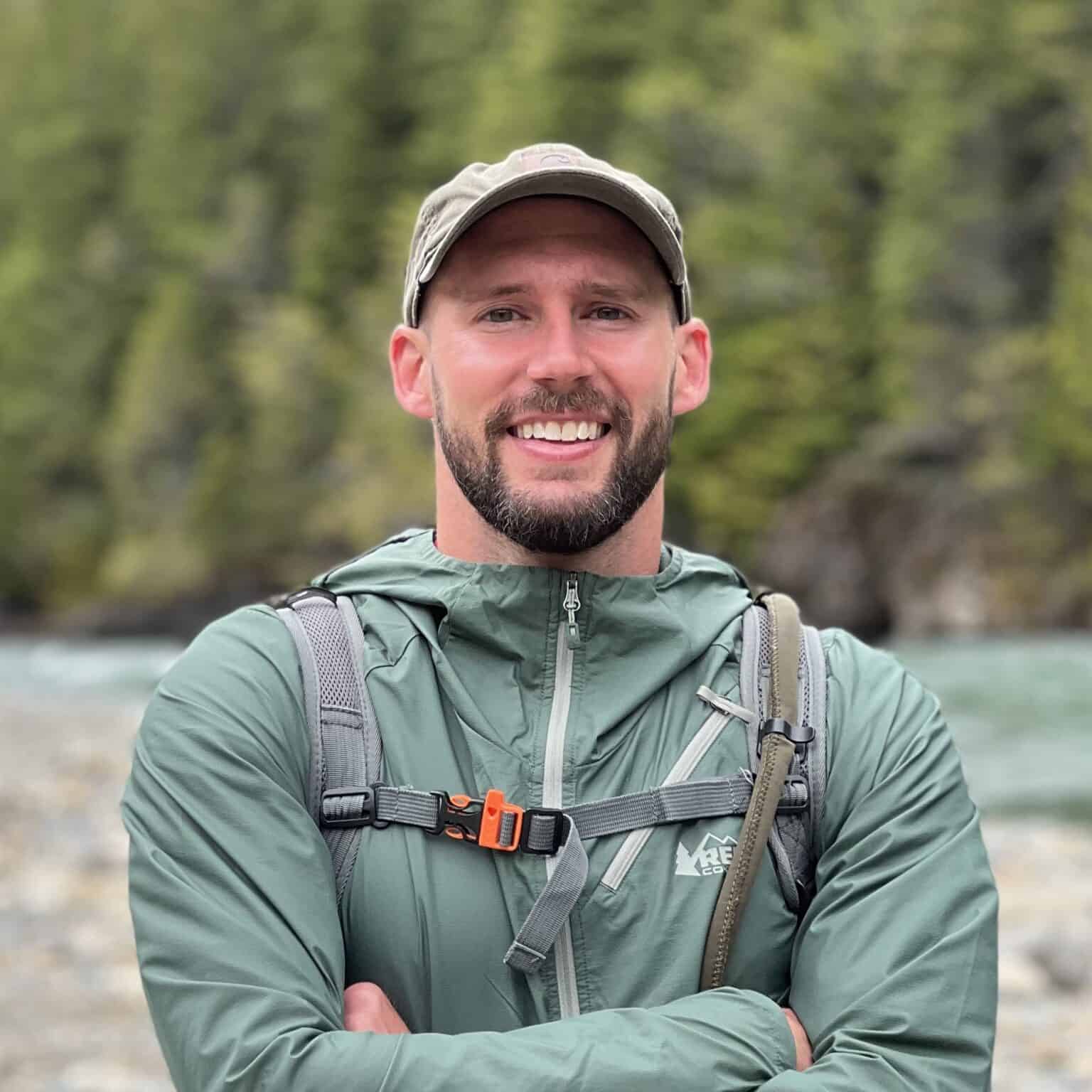 Beautiful man in outdoor adventure gear standing by river with lush green forest background.
