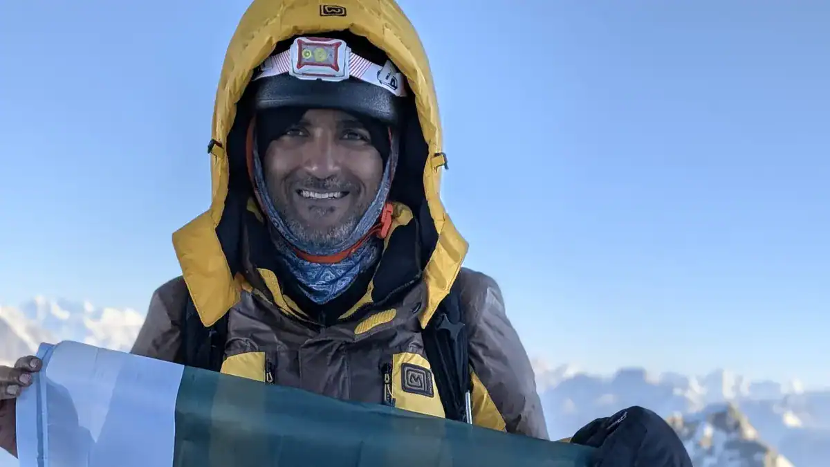 High-altitude mountaineer with winter gear holding a flag, smiling against a snowy mountain landscape, demonstrating endurance and training for extreme environments.