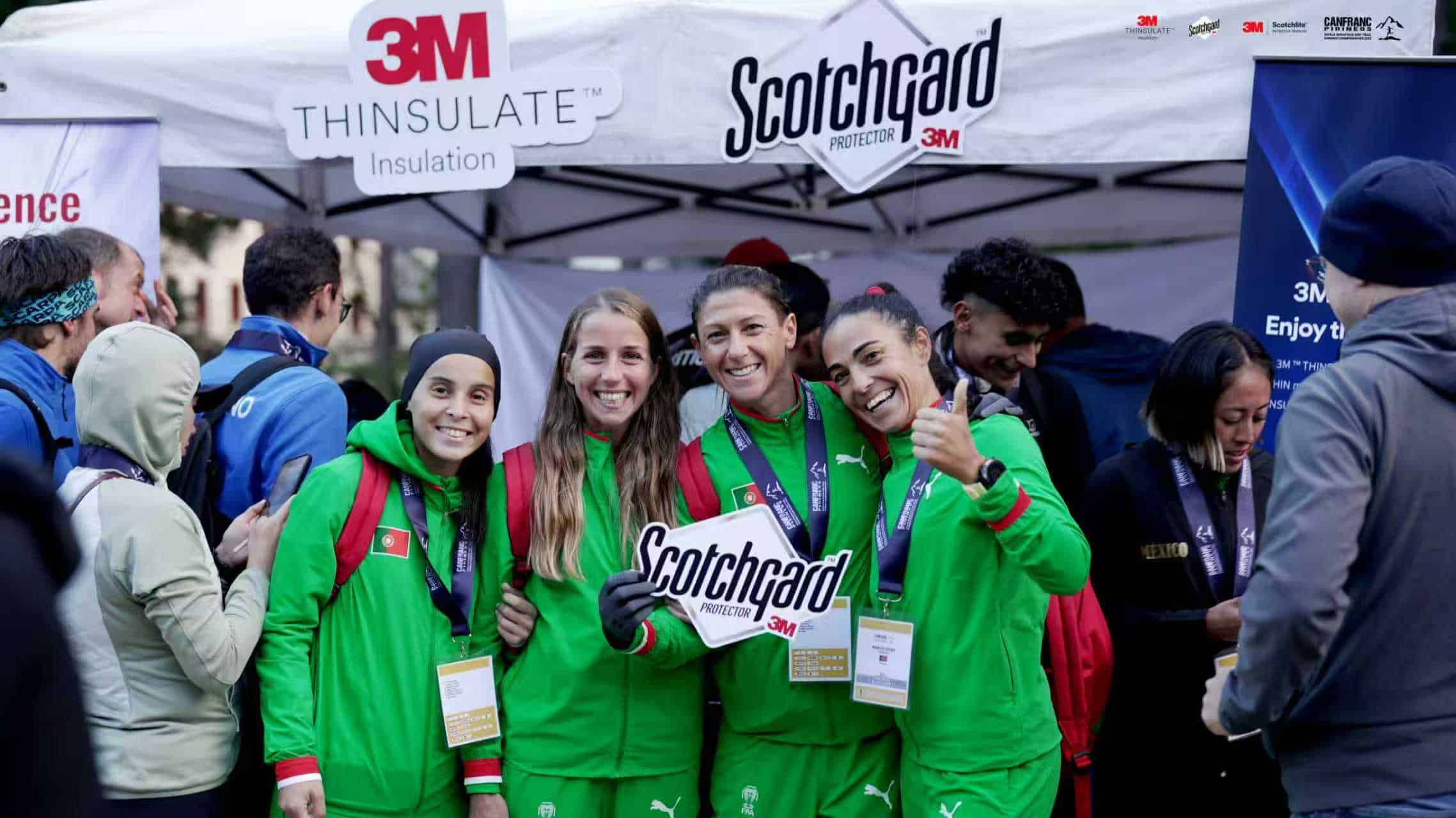 Happy female runners holding a Scotchgard sign at a marathon event, wearing green sports outfits with medals, surrounded by runners and spectators, promoting running sponsorship and athletic branding.
