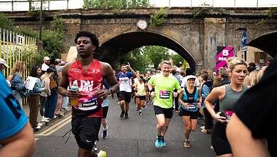 Runners participating in the Hackney Half Marathon, supporting the Single Homeless Project charity, with spectators and a historic bridge in the background.
