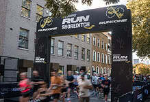 A detailed view of the starting line at the Shoreditch Run event, with runners in motion beneath a large black archway featuring event branding and sponsor logos, set against a backdrop of urban build.