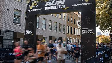 A detailed view of the starting line at the Shoreditch Run event, with runners in motion beneath a large black archway featuring event branding and sponsor logos, set against a backdrop of urban build.