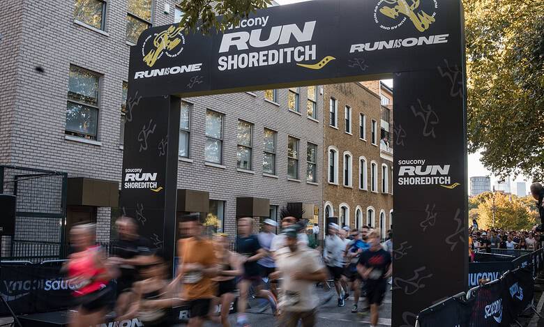 A detailed view of the starting line at the Shoreditch Run event, with runners in motion beneath a large black archway featuring event branding and sponsor logos, set against a backdrop of urban build.