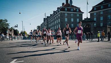 Running race with multiple athletes competing on a city street during daylight, with spectators and historic buildings in the background.
