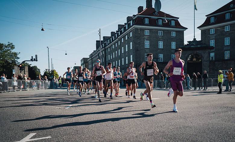 Running race with multiple athletes competing on a city street during daylight, with spectators and historic buildings in the background.