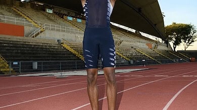 Running athlete smiling on a track at an outdoor stadium, wearing sports gear, ready for training or competition, with a clear blue sky and stadium seating in the background.