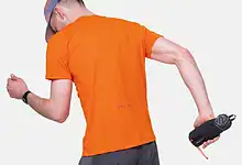 Close-up of a runner holding a Ronhill SoftFlask with Sleeve, wearing an orange shirt and grey shorts, preparing for a run.