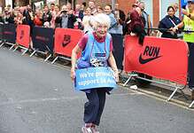 Elderly woman with white hair running in a marathon, holding a blue sign supporting St Ann's, with cheering crowd and race barriers in the background.