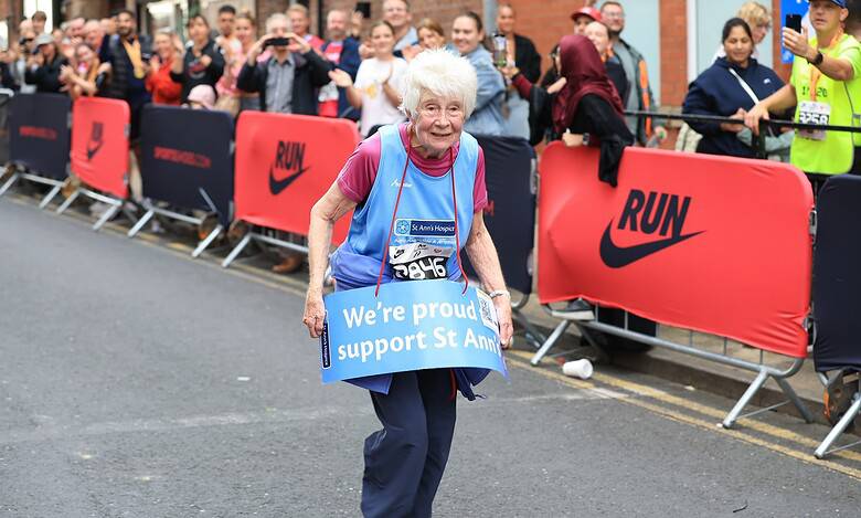 Elderly woman with white hair running in a marathon, holding a blue sign supporting St Ann's, with cheering crowd and race barriers in the background.
