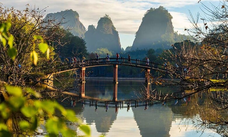 Beautiful river landscape with a wooden bridge, lush greenery, and towering karst mountains in the background, ideal for outdoor and travel enthusiasts.