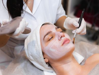 Beautician with a brush applies a white moisturizing mask to the face of a young girl client in a spa beauty salon
