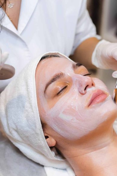 Beautician with a brush applies a white moisturizing mask to the face of a young girl client in a spa beauty salon