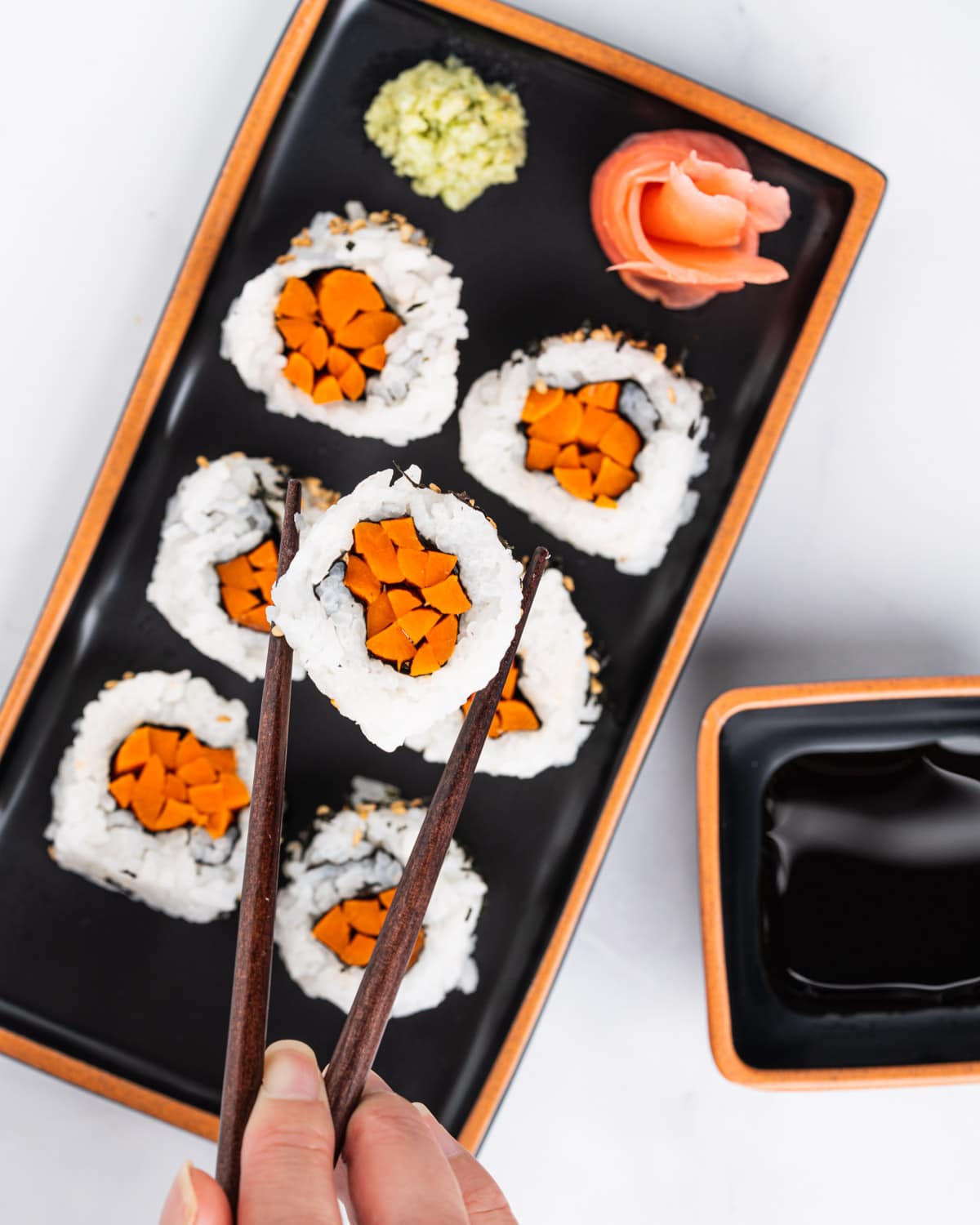 Chopsticks holding up one gobo sushi with a plate of gobo sushi rolls, wasabi and pickled ginger in the background.