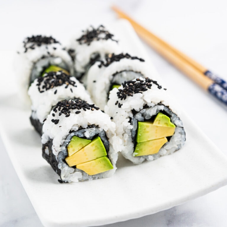 Sushi roll stuffed with creamy avocado standing up on a plate topped with black sesame seeds.