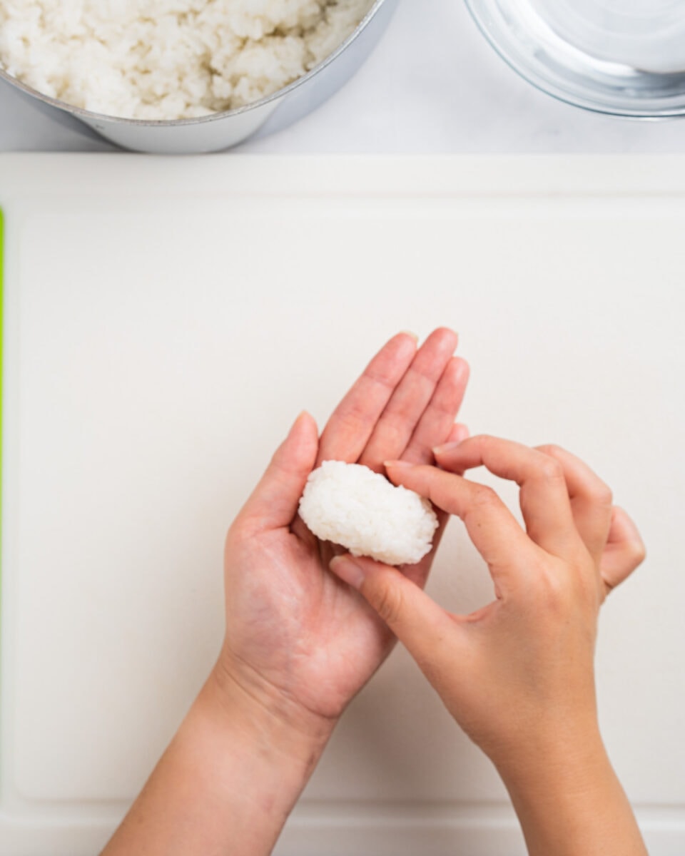 Hands shaping a small mound of rice. Rice and water bowl on the side.