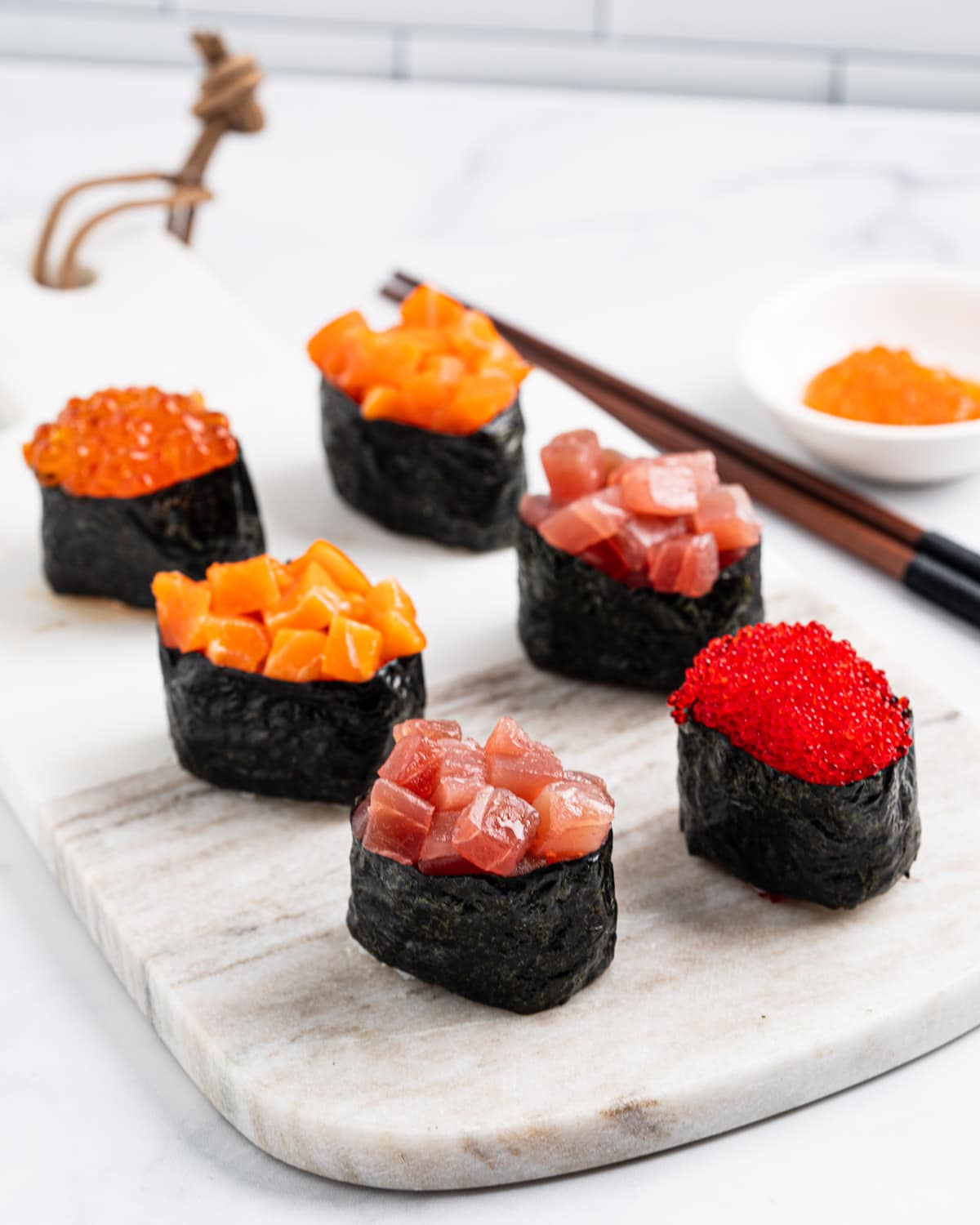 side angle of 6 gunkan on a marble serving board: 1 tobiko gunkan, 2 tuna gunkans, 2 salmon gunkans and 1 ikura gunkan.