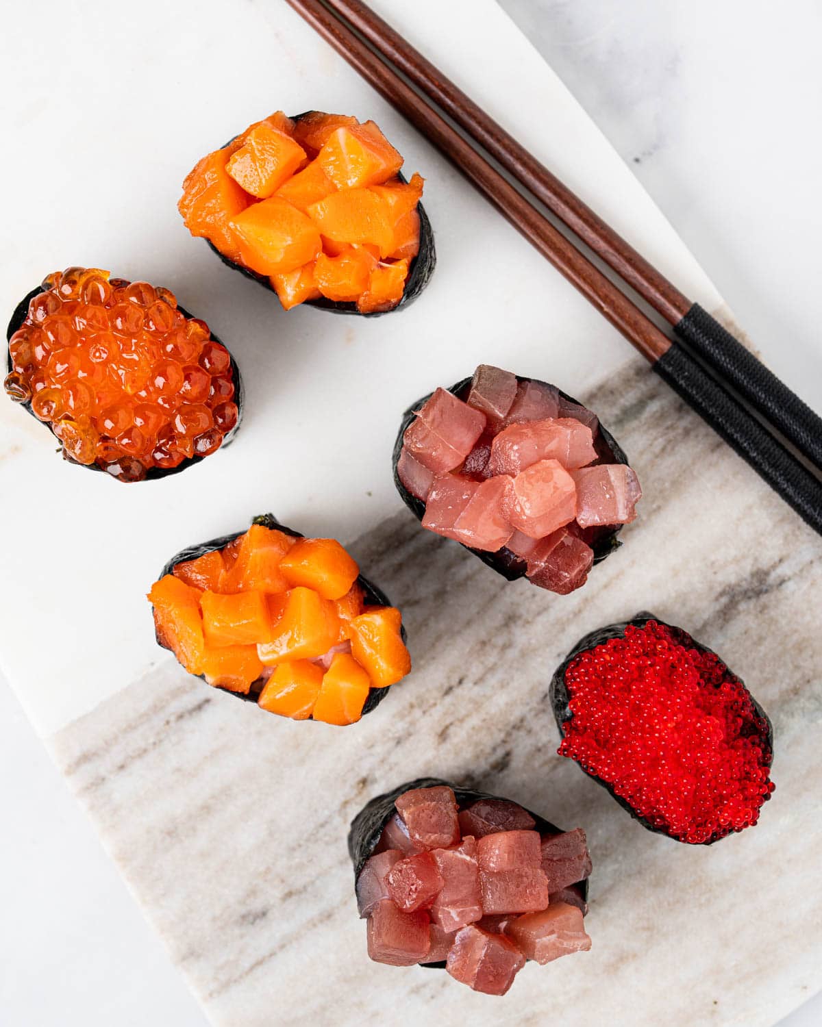 top down angle of 6 gunkan on a marble serving board: 1 tabiko gunkan, 2 tuna gunkans, 2 salmon gunkans and 1 ikura gunkan.